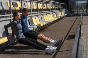 Two people are doing triceps dips on a low barrier beside stadium seating, exercising outdoors at a sports track.